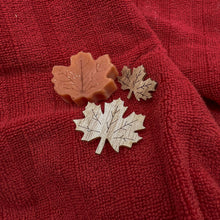 Load image into Gallery viewer, Two wooden maple leaves and one small decorative soap leaf on red fabric.