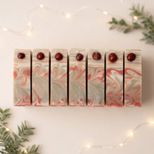 Charger l'image dans la galerie, Seven Christmas soap bars in a row, each topped with a red jasper stone above red, cream, and pale green swirls.