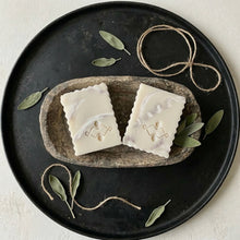 Load image into Gallery viewer, Two pieces of white soap with a Woods and Mosses logo on a stone tray with green leaves and twine on a black plate.