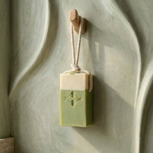 Load image into Gallery viewer, Matcha latte soap on a rope bar hanging from a wooden hook, showing the front stamp and braided cord.