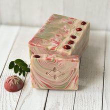 Load image into Gallery viewer, Handcrafted Christmas soap bars with red, cream, and green swirls, sparkling mica on top, and glossy red embeds. The front bar features a stamped geometric logo, giving a festive peppermint-candy look. Natural cold process holiday artisan soap.