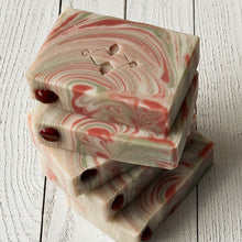 Load image into Gallery viewer, Stack of handmade Christmas soap bars with cream, red, and green swirls in circular patterns, stamped with a geometric logo and decorated with glossy red embeds. The marbled design and shimmering mica give a festive candy-cane appearance.