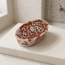 Load image into Gallery viewer, Oval Moroccan clay soap with carved floral top, white marbling, and warm terracotta speckles