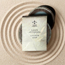 Load image into Gallery viewer, Round artisan soap bar wrapped in Woods and Mosses branded packaging, with blue and brown layered swirl bars visible behind it