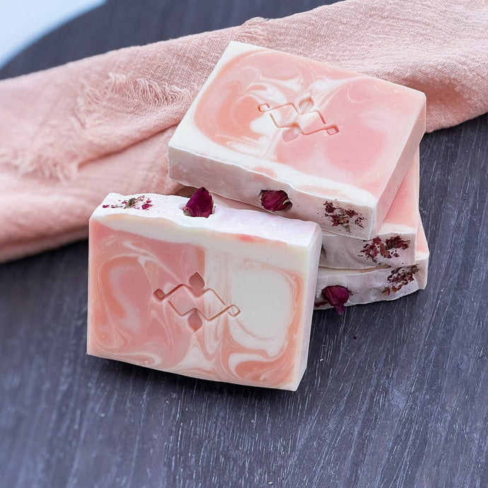 Artisan rose soap bars with a pink and cream swirl pattern and geometric Woods and Mosses stamp, topped with dried rosebud and petal for a floral skincare aesthetic.