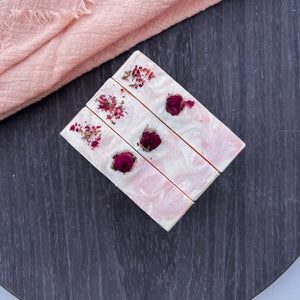 Top view of three floral soap bars with dried rosebuds and petals embedded on a creamy pink surface, featuring gentle marbled swirl and botanical decor.