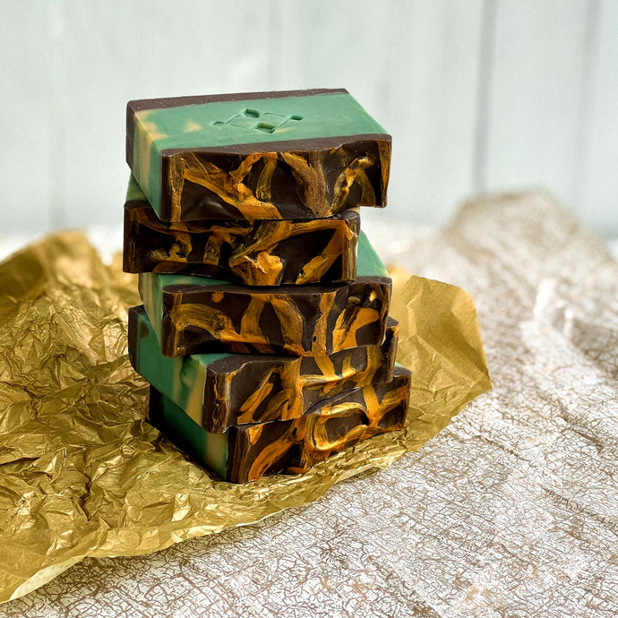Artisan soap bars stacked vertically, each with a green base, dark cocoa top, and golden swirl. Cold process handmade soap with embossed mark, ideal for natural skincare and gift sets.