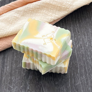 Three pastel artisan soap bars stacked diagonally, with marbled swirls of pink, yellow, green, and white, placed on a dark round background with soft fabric.