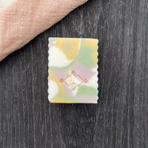 Single rectangular artisan soap bar with soft pastel swirls and textured edges, centered on a dark surface with a peach linen fabric in the corner.