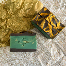 Charger l&#39;image dans la galerie, Three handmade soap bars displayed flat, one showing a green face with an embossed logo, the other showing a top view with bold golden swirls on a dark chocolate surface. Artisan cold process design for natural skincare and gifting.