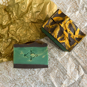 Three handmade soap bars displayed flat, one showing a green face with an embossed logo, the other showing a top view with bold golden swirls on a dark chocolate surface. Artisan cold process design for natural skincare and gifting.