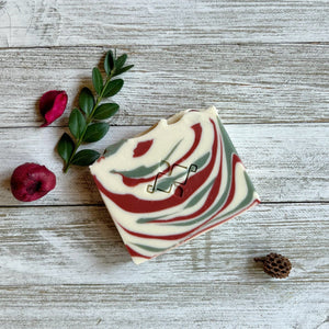 A square soap bar with the Woods and Mosses company logo in the middle. It is white with red and green swirls. 