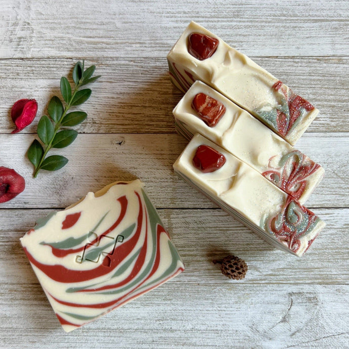 A square soap bar with the Woods and Mosses company logo in the middle. It is white with red and green swirls. The top is textured and decorated with red gemstones and glitter.