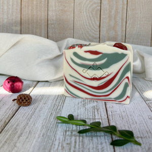 A square soap bar with the Woods and Mosses company logo in the middle. It is white with red and green swirls. The top is decorated with red gemstones.